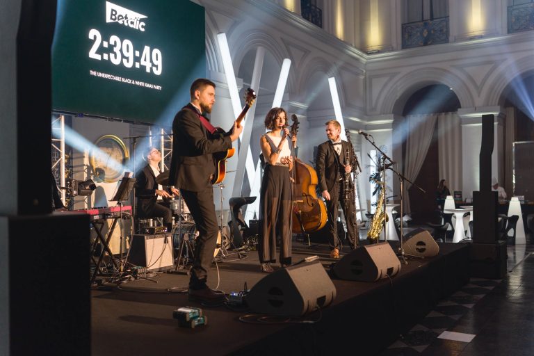 Un groupe de jazz se produit sur une scène dans une salle élégante aux hauts plafonds. Lors de la Soirée annuelle du Groupe Betclic, une femme chante dans un micro, accompagnée de musiciens jouant de la guitare, du saxophone, de la contrebasse et de la batterie. Une horloge numérique affiche 2:39:49. L'ambiance est sophistiquée avec un éclairage dramatique et des gens assis en arrière-plan.