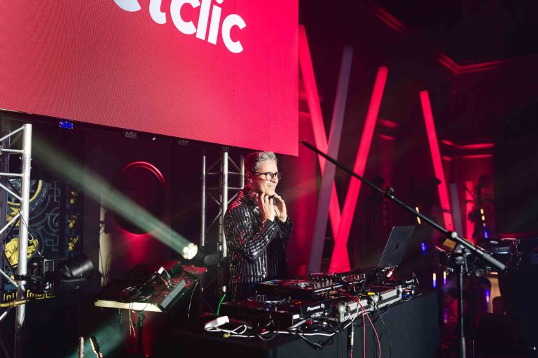 Un DJ aux cheveux courts et aux lunettes, vêtu d'une veste rayée noire et argentée, se tient derrière une console de mixage sur une scène de la Soirée annuelle du Groupe Betclic. Il a les mains jointes comme s'il applaudissait. Derrière lui, un écran numérique éclairé en rouge affiche « clic », et la scène est équipée de divers équipements d'éclairage et de sonorisation dans une salle sombre.