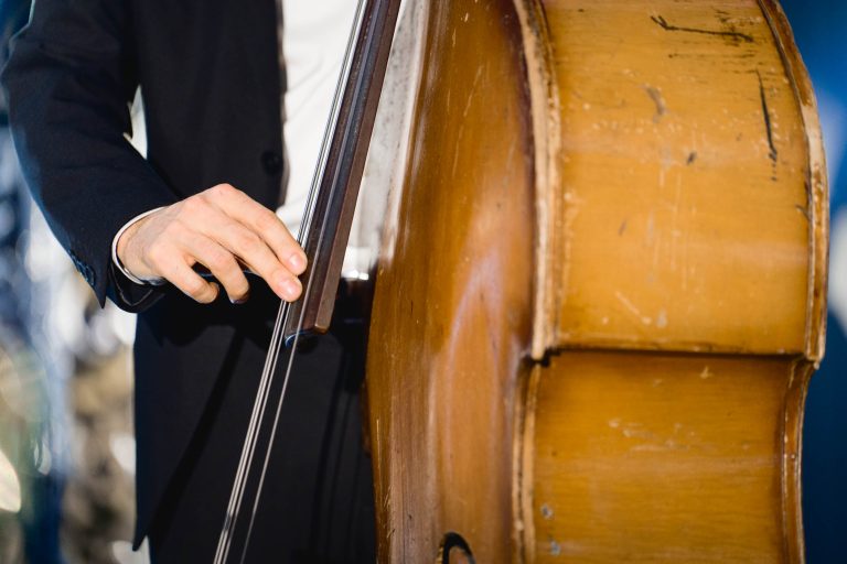 Une personne en costume noir joue de la contrebasse verticale à la Soirée Annuelle du Groupe Betclic. L'image met en valeur la main gauche de la personne qui pince les cordes et le corps de l'instrument. La contrebasse a une finition en bois naturel avec des marques d'usure visibles, indiquant une utilisation intensive. L'arrière-plan est flou.