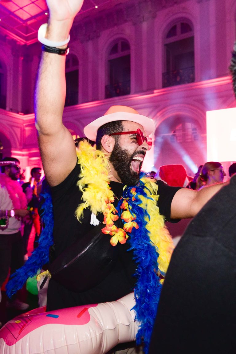 Un homme festif et barbu, coiffé d'un chapeau blanc, de lunettes de soleil rouges et d'un boa en plumes colorées, lève le bras avec énergie lors de la Soirée annuelle du Groupe Betclic. L'arrière-plan présente un bâtiment décoré avec des fenêtres cintrées baignées d'un éclairage rose et violet, et d'autres fêtards sont partiellement visibles.