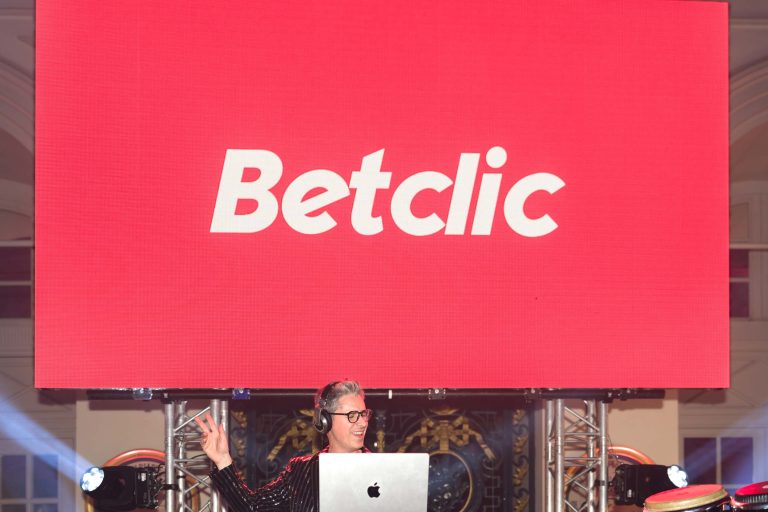 Un DJ portant des écouteurs se tient debout derrière un ordinateur portable, souriant et faisant des gestes de paix. Au-dessus de lui, un grand écran rouge affiche le mot « Betclic » en texte blanc. L'arrière-plan comprend des lumières et des équipements de scène, capturant l'atmosphère vibrante de la Soirée Annuelle du Groupe Betclic.