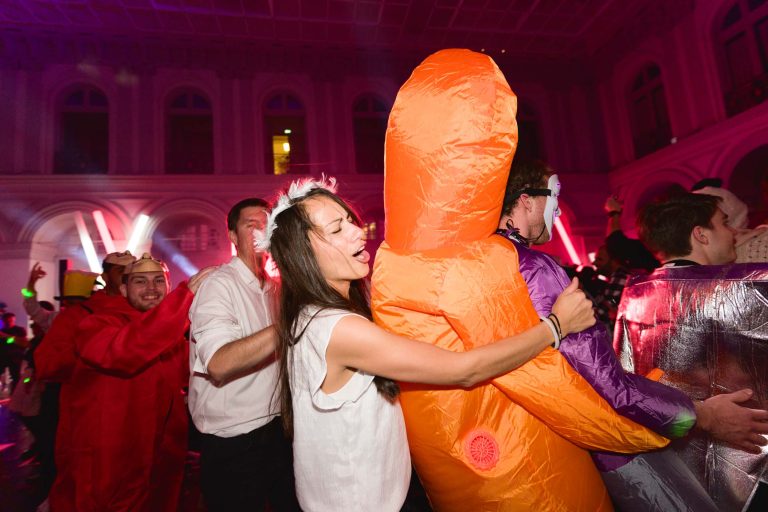 Une scène de fête animée avec des gens qui dansent. Une femme en haut blanc sourit en dansant avec quelqu'un en costume gonflable orange. L'arrière-plan est rempli d'autres fêtards en tenues festives colorées sous un éclairage violet et rouge vibrant, célébrant la Soirée annuelle du Groupe Betclic.