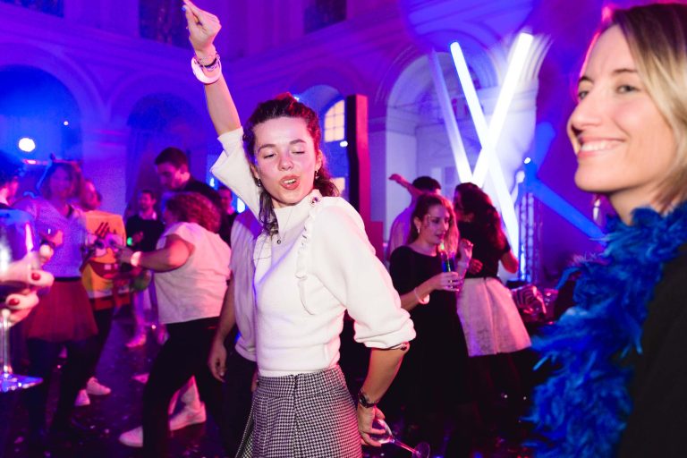 Lors de la Soirée annuelle du Groupe Betclic, une scène de fête animée se déroule dans une salle spacieuse aux hauts plafonds et aux fenêtres cintrées. Une femme au premier plan danse avec énergie, vêtue d'un haut blanc et d'une jupe à carreaux. Une autre femme à droite, souriante, est vêtue de vêtements sombres et d'un boa en plumes bleues. Des lumières colorées illuminent l'arrière-plan.