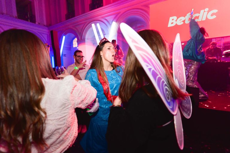 Une scène de fête animée dans un club au plafond haut et aux fenêtres cintrées. Plusieurs personnes sont habillées de tenues colorées ; l'une porte une robe bleue et un diadème, une autre a des ailes de papillon. Un grand écran en arrière-plan affiche « Soirée annuelle Betclic Group ». L'ambiance est animée, avec des lumières vives et de la musique.