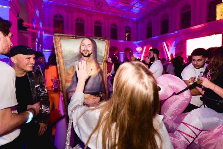 Une soirée annuelle animée du groupe Betclic avec des personnes déguisées. Une femme au centre interagit avec une personne déguisée en Mona Lisa. La salle est éclairée par un éclairage violet vif et un grand plafond. Les fêtards qui les entourent sont habillés de manière décontractée et semblent apprécier l'événement.