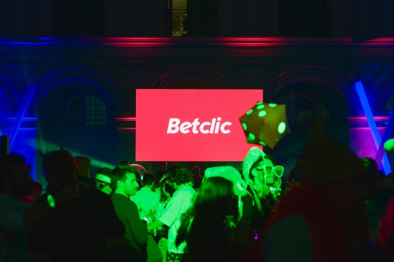 Un grand écran rouge affichant le mot « Betclic » en caractères blancs est bien en évidence au centre de l'image de la Soirée annuelle du Groupe Betclic. L'événement est doté d'un éclairage vibrant et multicolore et d'une piste de danse bondée, où les gens s'amusent, plusieurs d'entre eux étant parés de costumes et d'accessoires amusants.