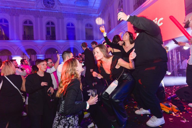 Lors de la soirée annuelle animée du groupe Betclic, les gens dansent et s'amusent au milieu de lumières colorées. Certains sont debout sur une estrade, tenant des boissons et applaudissant. L'arrière-plan présente l'intérieur d'un bâtiment à plusieurs étages décoré et des confettis sont dispersés sur le sol. Tout le monde est habillé en tenue de fête.