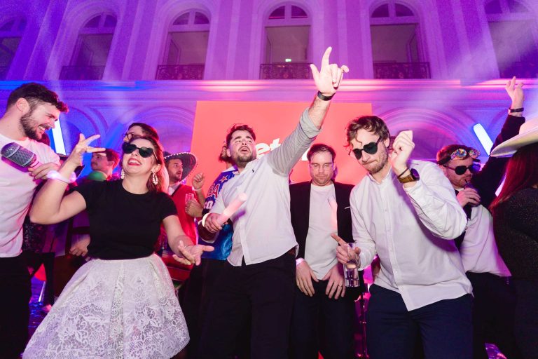 Un groupe de personnes dansent et chantent sur scène lors de la soirée annuelle animée du Groupe Betclic. L'ambiance est renforcée par un éclairage coloré, à dominante rose et violet. Certains participants portent des costumes et des accessoires comme des lunettes de soleil et des chapeaux, ajoutant à l'ambiance festive. En arrière-plan, un grand écran rouge est visible.