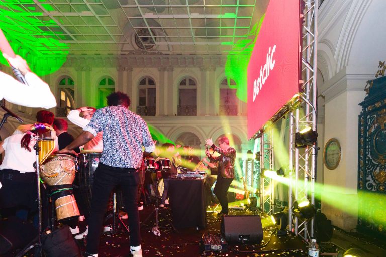 Un concert animé en salle à la Soirée annuelle du Groupe Betclic met en vedette un DJ et des musiciens sur scène. Le groupe joue de divers instruments sous un éclairage vert et jaune vibrant. Un plafond orné et de grandes fenêtres cintrées sont visibles en arrière-plan, avec un grand écran rouge affichant du texte à côté de la scène.