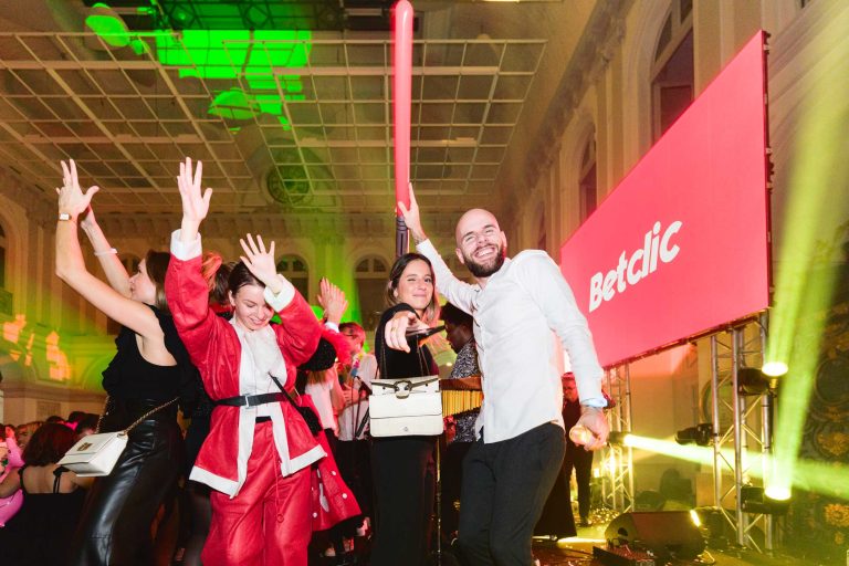 Les gens dansent joyeusement lors d'une soirée annuelle animée du groupe Betclic. Une femme en costume de Père Noël tient un bâton lumineux et lève le bras. Un homme à côté d'elle sourit et lève également le bras. L'arrière-plan est composé de lumières vives et colorées et d'un grand écran affichant le mot « Betclic ». L'ambiance est festive et énergique.