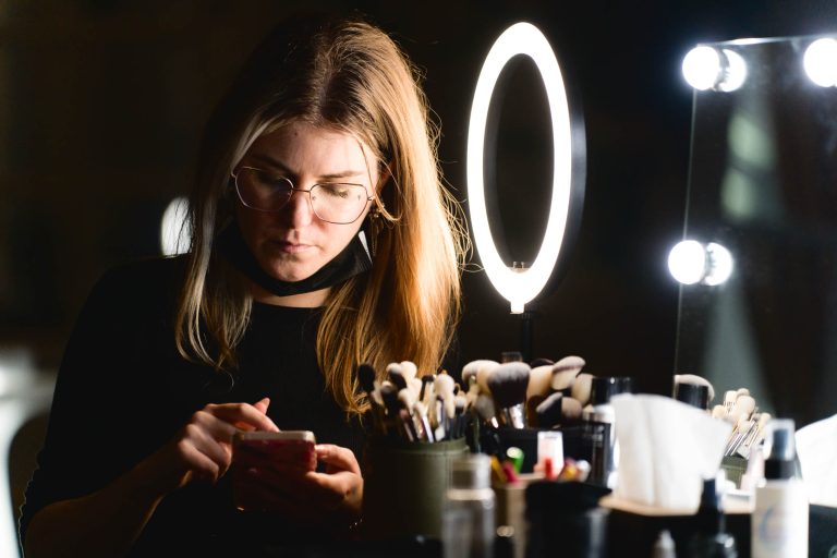 Une femme aux cheveux longs et aux lunettes utilise son smartphone tout en étant assise devant une coiffeuse avec un anneau lumineux et divers outils de maquillage. Elle apparaît concentrée, entourée de pinceaux, d'articles de maquillage et d'un miroir avec des lumières rondes, se préparant pour la soirée annuelle du groupe Betclic. Le décor est faiblement éclairé, mettant en valeur la lueur de l'anneau lumineux.