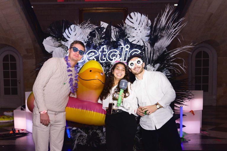 Trois personnes posent pour une photo lors de la Soirée annuelle du Groupe Betclic. Toutes sourient. La personne de gauche porte des lunettes de soleil, une chemise blanche et un collier de fleurs violet. La personne du milieu porte une tenue noire et tient un verre. La personne de droite est habillée en blanc, avec des lunettes de soleil originales. Derrière elles se trouvent un canard gonflable coloré et des décorations à thème tropical.