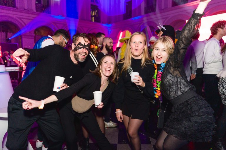 Un groupe de quatre personnes posent joyeusement lors de la soirée annuelle animée du groupe Betclic. Ils sont dans un décor festif avec un éclairage coloré. Une personne porte un masque de mascarade et une autre a un collier de fleurs vibrant autour du cou. Ils semblent tous s'amuser, sourire et tenir des tasses.