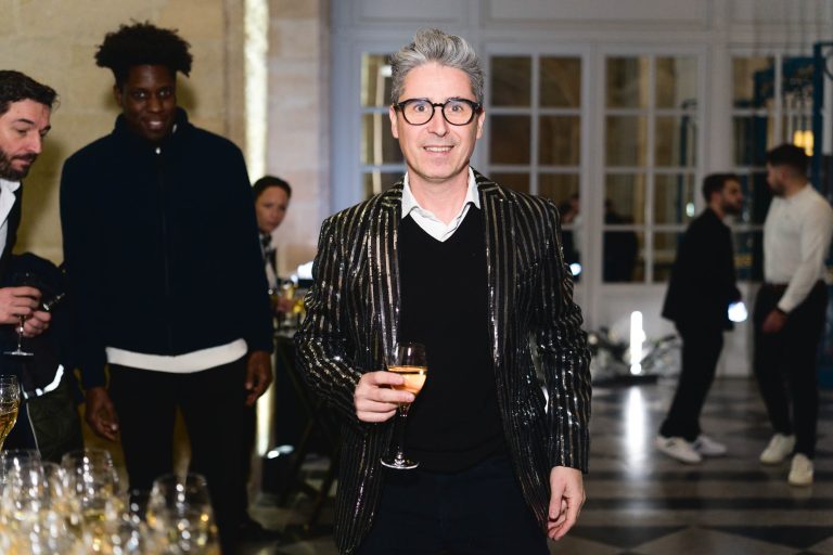 Un homme aux cheveux gris et aux lunettes, vêtu d'une veste noire brillante sur une chemise noire et un col blanc, tient une coupe de champagne à la soirée annuelle du Groupe Betclic. Il se tient dans une pièce aux grandes fenêtres et au sol en damier, tandis que plusieurs autres personnes en arrière-plan discutent.