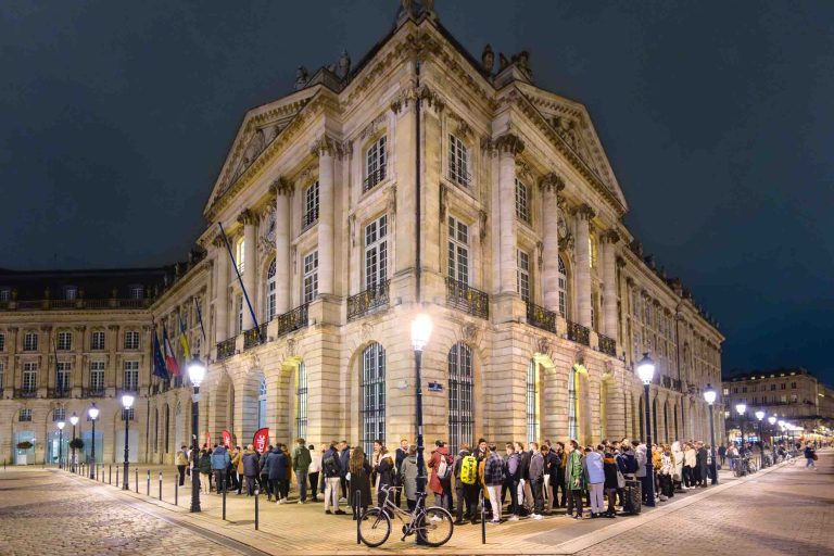 Une foule de gens fait la queue pour la Soirée annuelle du Groupe Betclic, devant un bâtiment historique décoré et bien éclairé, la nuit. Le bâtiment est doté de grandes fenêtres, de balcons et de pierres décoratives. Plusieurs lampadaires éclairent la scène et des vélos sont garés à proximité. Le ciel est sombre et la zone est pavée de pavés.