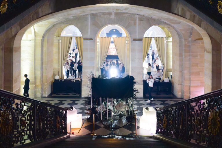 Un intérieur grandiose avec un large escalier mène à un hall élaboré aux fenêtres en arc, le cadre de la soirée annuelle du groupe Betclic. Les invités, vêtus de tenues de soirée, se mêlent et discutent en groupes. Un éclairage doux met en valeur le décor élégant, notamment les balustrades en fer forgé et le sol à carreaux. De hauts rideaux encadrent les portes en arc.