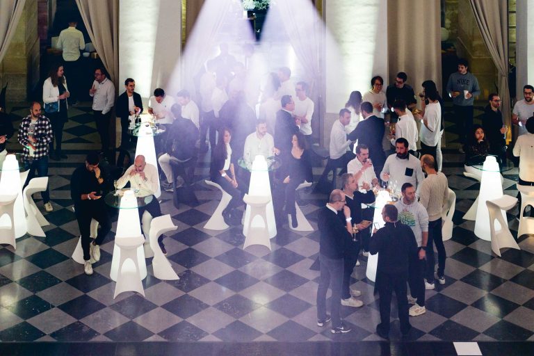 Lors de la Soirée annuelle du Groupe Betclic, un grand rassemblement se déroule dans un espace intérieur élégant au sol à carreaux et aux grands rideaux. Autour de tables blanches modernes et lumineuses, les invités discutent et échangent. Un éclairage doux avec des poutres met en valeur certains espaces, renforçant l'ambiance élégante.