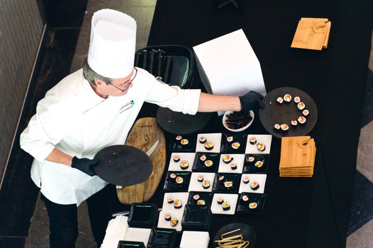 Lors de la soirée annuelle du groupe Betclic, un chef vêtu d'un uniforme blanc et d'un grand chapeau blanc dispose soigneusement des rouleaux de sushi sur des plateaux de service noirs. La table est dressée avec divers plats carrés blancs et noirs, des planches en bois et des serviettes blanches. Le chef utilise des pinces et des gants noirs pour manipuler la nourriture.