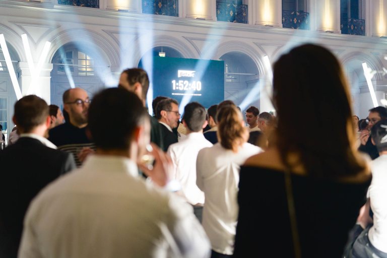 Une foule nombreuse se rassemble à l'intérieur pour un événement où les gens, habillés en tenue semi-formelle, se mêlent et discutent. La salle est illuminée par des colonnes lumineuses et des projecteurs suspendus. En arrière-plan, une grande horloge numérique de compte à rebours affichant « 1:52:40 » donne le ton à la Soirée annuelle du Groupe Betclic.