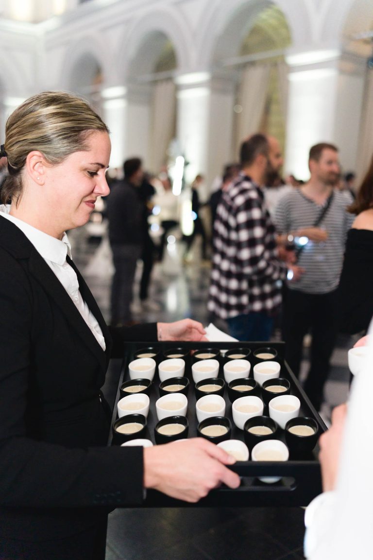 Un serveur en uniforme noir tient un plateau rempli de petites tasses blanches lors de la Soirée Annuelle du Groupe Betclic. Le lieu est agrémenté de hautes arches et de colonnes blanches. À l'arrière-plan, les participants, certains en tenue décontractée, discutent et tiennent des assiettes ou des tasses au milieu de la foule qui se mêle.