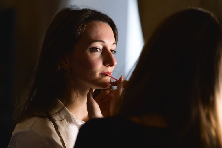 Une femme aux cheveux noirs mi-longs se fait maquiller. Elle regarde légèrement sur le côté tandis qu'une autre personne, presque hors cadre avec seulement ses mains visibles, applique du rouge à lèvres sur ses lèvres. L'éclairage est doux avec des ombres mettant en valeur ses traits du visage, se préparant pour la soirée annuelle du groupe Betclic.