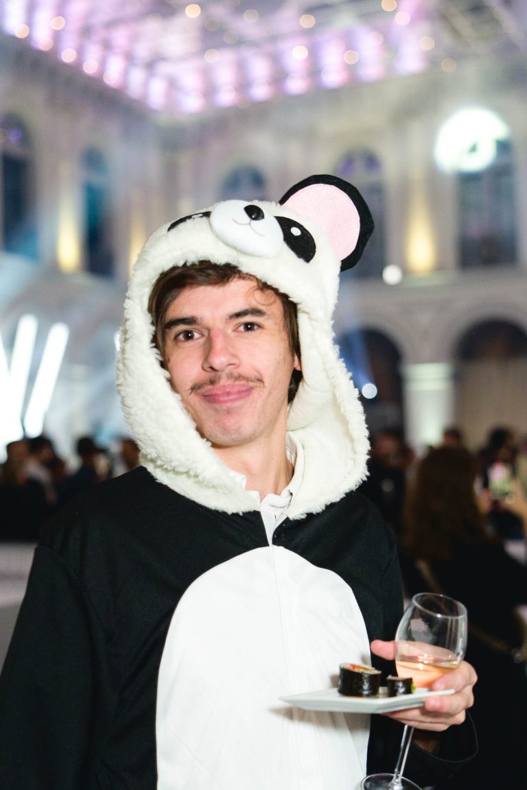 Une personne portant un costume de panda avec une capuche se tient à l'intérieur lors de la Soirée annuelle du Groupe Betclic, où les fenêtres cintrées sont illuminées de lumières violettes. Elle tient un verre de vin blanc dans une main et une petite assiette de nourriture dans l'autre, souriant à la caméra. L'arrière-plan montre d'autres participants en train de socialiser.