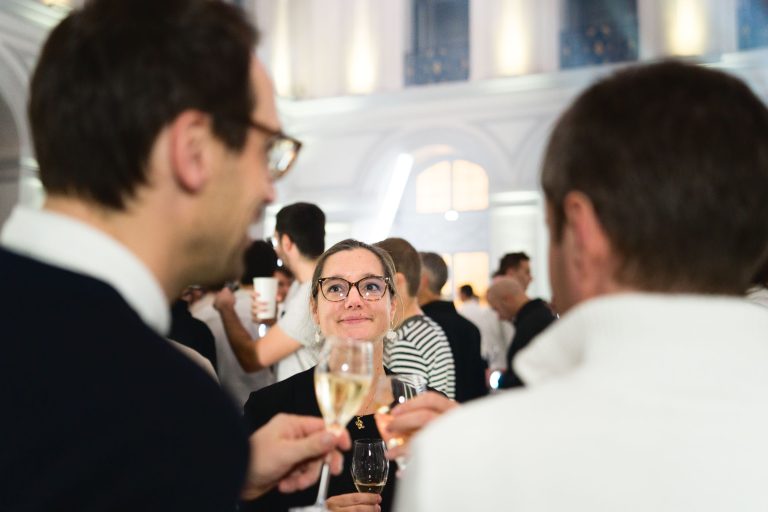 Lors de la Soirée annuelle du Groupe Betclic, un groupe de personnes socialise lors d'un événement intérieur. Une femme portant des lunettes, au centre, tient un verre de champagne et sourit en regardant deux individus au premier plan qui tiennent également des boissons. L'arrière-plan présente un décor élégant et bien éclairé avec d'autres invités qui se mêlent.