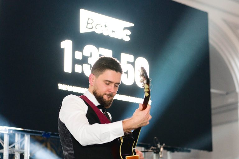 Un homme en chemise blanche, gilet noir et cravate rouge joue de la guitare électrique sur scène lors de la soirée annuelle du groupe Betclic. Derrière lui, un grand compte à rebours numérique affiche « 1:37:50 » et le mot « Betclic ». Des lumières vives illuminent la scène, mettant en valeur l'expression concentrée de l'interprète.