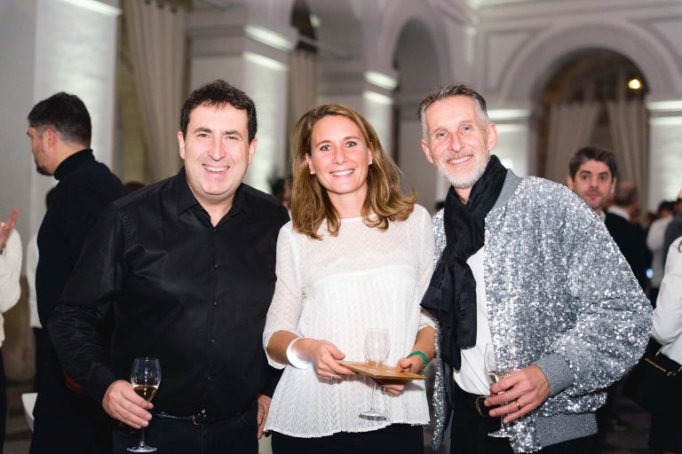 Trois personnes posent ensemble lors de la soirée annuelle du Groupe Betclic. L'homme à gauche porte une chemise noire et tient un verre, la femme au centre porte un chemisier blanc avec un verre et une petite assiette, et l'homme à droite porte une veste argentée étincelante et une écharpe noire, tenant également un verre. Les gens et l'architecture en arc sont en arrière-plan.