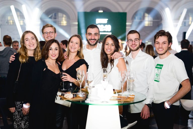 Un groupe de huit jeunes adultes, habillés en tenue semi-formelle, se rassemblent autour d'une table haute ornée de verres à vin vides lors de la Soirée annuelle du Groupe Betclic. Ils se tiennent debout dans un espace événementiel intérieur bien éclairé avec des plafonniers blancs brillants et un écran d'affichage vert en arrière-plan, tous souriant à la caméra.