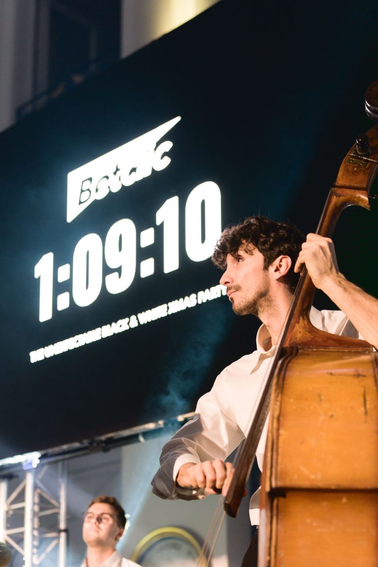 Un musicien aux cheveux bouclés et portant une chemise blanche joue de la contrebasse sur scène. En arrière-plan, un grand chronomètre numérique affiche 1:09:10. Au-dessus du chronomètre se trouve le mot « Betclic » et en dessous, le texte « The Unofficial Black & White Xmas Party ». Un autre musicien est partiellement visible lors de cette Soirée annuelle du Groupe Betclic.