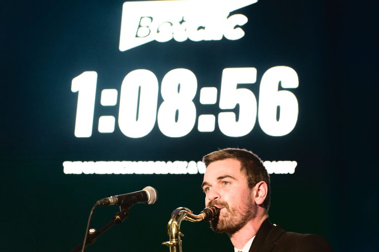 Un homme aux cheveux courts et à la barbe barbue joue du saxophone au micro. Derrière lui, un grand chronomètre numérique indique « 1:08:56 », une partie d'un panneau sur lequel figure « Betclic ». La scène se déroule sur un fond sombre lors de la Soirée annuelle du Groupe Betclic.