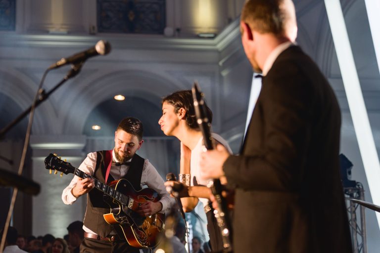 Trois musiciens se produisent sur scène lors de la Soirée annuelle du Groupe Betclic, habillés en tenue de soirée. Le guitariste de gauche joue avec énergie, tandis que celui du centre semble chanter. Le troisième musicien, à droite, tient une guitare et observe. Ils se trouvent dans une salle bien éclairée avec un public attentif en arrière-plan.