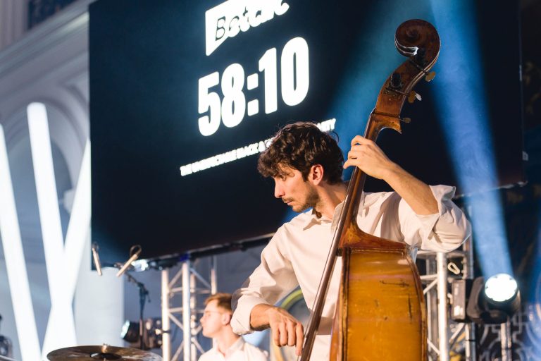 Un musicien en chemise blanche joue de la contrebasse sur scène lors de la Soirée annuelle du Groupe Betclic. Derrière lui, un grand écran affiche le mot « Botak » et un chronomètre indiquant « 58:10 ». Des lumières de scène et du matériel musical sont visibles, et une autre personne est partiellement vue en train de jouer de la batterie en arrière-plan.