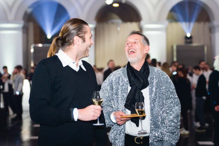 Deux hommes en tenue semi-formelle tiennent des verres de vin à la soirée annuelle animée du groupe Betclic. Ils rient et discutent. L'homme de gauche porte un pull noir avec une chemise à col blanc en dessous, et l'homme de droite porte une veste argentée scintillante avec une écharpe noire. Des personnes et des lumières sont visibles dans l'arrière-plan flou.