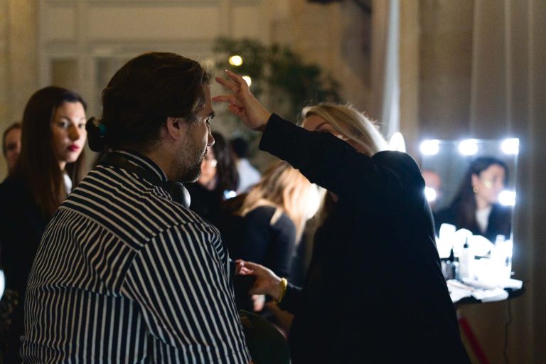 Une maquilleuse maquille un homme barbu, vêtu d'une chemise rayée noire et blanche. La salle, doucement éclairée, bourdonne d'activité pendant la soirée annuelle du Groupe Betclic, avec d'autres personnes en arrière-plan, certaines assises, d'autres debout. Un miroir éclairé et divers produits de maquillage sont visibles sur une table à droite.