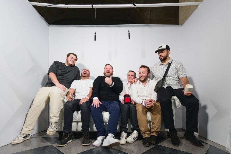 Six hommes sont assis tranquillement sur un canapé blanc dans une pièce aux murs blancs et au sol à carreaux pendant la soirée annuelle du groupe Betclic. Ils rient et sourient, la plupart vêtus de vêtements décontractés et l'un d'eux tenant une tasse. L'atmosphère générale semble détendue et amicale. Deux microphones sont placés au plafond.