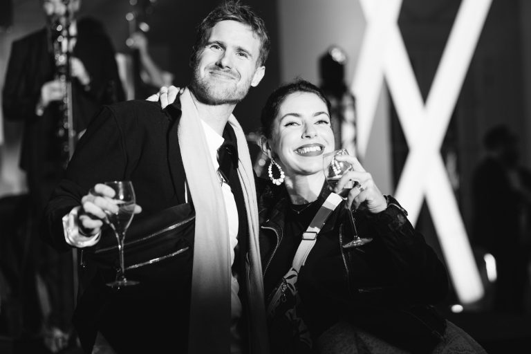 Un homme et une femme sourient tout en tenant des verres à vin lors d'une élégante soirée annuelle organisée par le groupe Betclic. L'image en noir et blanc les montre habillés de manière formelle, l'homme portant un costume et une écharpe, et la femme ornée de grandes boucles d'oreilles créoles et d'un ensemble sombre. L'arrière-plan présente des lumières floues et d'autres participants.