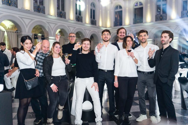 Un groupe de dix personnes se rassemblent à l'intérieur, souriant et levant leurs verres pour célébrer leur anniversaire. Le lieu élégant avec ses hauts plafonds et ses fenêtres cintrées donne le ton à l'ambiance animée de la Soirée Annuel Betclic Group, avec des invités en tenue décontractée ou formelle.