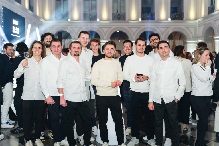 Un groupe de douze personnes, en majorité des hommes, se rassemble dans une salle spacieuse avec de hauts plafonds et des arcades lors de la soirée annuelle du groupe Betclic. Ils sont vêtus de chemises et de pulls blancs décontractés, certains portant des pantalons noirs. Des gens à l'arrière-plan discutent. L'atmosphère semble festive et animée.