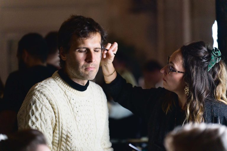 Un homme aux yeux clos est assis tandis qu'une femme, portant des lunettes et une pince à cheveux, lui maquille le visage. Vêtu d'un épais pull torsadé de couleur crème, il est assis au milieu de l'ambiance de la soirée annuelle du groupe Betclic, où les autres convives se mêlent dans un arrière-plan faiblement éclairé.