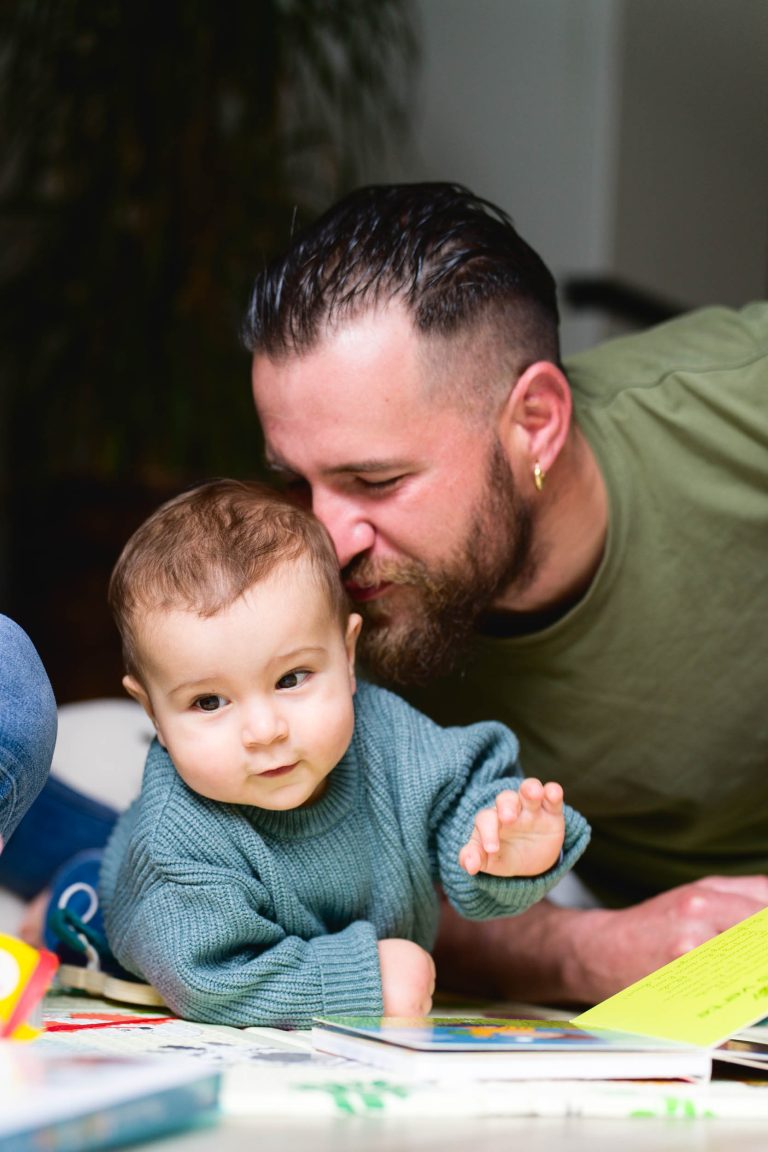 Un homme barbu aux cheveux courts et portant une boucle d'oreille se penche pour embrasser un bébé sur la tête. Le bébé, vêtu d'un pull en tricot bleu sarcelle, regarde vers l'avant avec une expression calme tout en tendant la main vers un livre ouvert sur la table. L'arrière-plan de cette séance photo de la famille Bordelaise est légèrement flou avec des plantes vertes visibles.