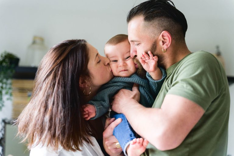 Sur une photo de la séance de la famille Bordelaise, une femme aux cheveux bruns portant une chemise blanche et un homme barbu portant une boucle d'oreille en or portant une chemise verte tiennent et embrassent affectueusement leur bébé. Vêtu d'un pull vert et d'un jean bleu, le bébé satisfait tend la main vers le visage de l'homme.