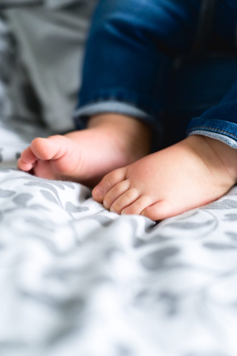 Gros plan sur les pieds nus d'un enfant posés sur une couverture à motifs gris et blanc. L'enfant porte un jean bleu et l'attention est portée sur la plante des pieds minuscules, aux orteils légèrement recourbés. L'arrière-plan, bien que flou, évoque l'atmosphère douce et douillette typique d'une séance photo de la famille Bordelaise.