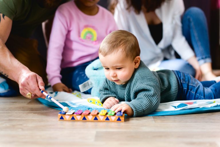 Un bébé en pull gris est allongé sur un tapis de jeu coloré posé sur un plancher en bois, tendant la main vers un xylophone joué par la main d'un adulte tenant un maillet bleu et blanc. Pendant cette séance photo de la famille Bordelaise, deux autres personnes, dont un enfant en pull rose, sont assises et observent en arrière-plan.