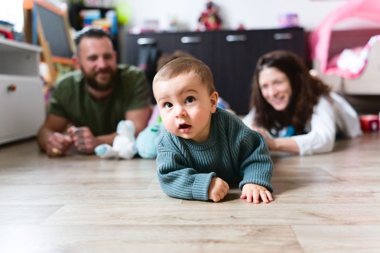 Un bébé en pull vert rampe sur un plancher en bois, l'air curieux. En arrière-plan, un homme barbu en chemise verte et une femme en haut blanc sont allongés sur le sol, souriant au bébé. La pièce est meublée de jouets et d'armoires, capturant une séance photo intime de la famille Bordelaise.