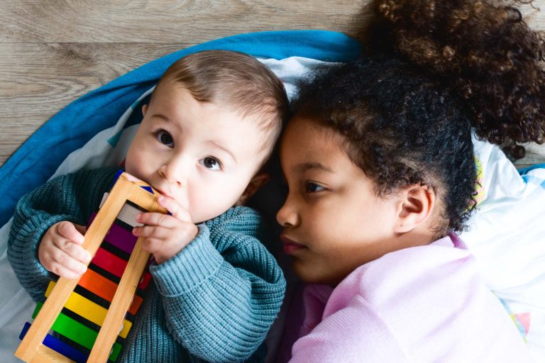 Un bébé aux cheveux courts, portant un pull vert, est allongé et tient un xylophone coloré, le regard tourné vers le haut. A côté du bébé, un enfant plus âgé aux cheveux bouclés, portant un haut rose, pose sa tête sur la même couverture, le regard tourné vers le côté. Tous deux sont sur un plancher en bois avec un tapis bleu en dessous pendant leur séance photo famille Bordelaise.