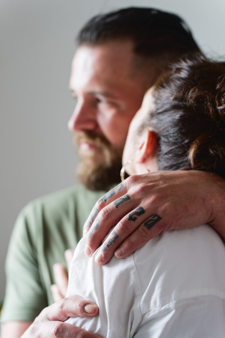 Un homme barbu enlace une femme pendant leur séance photo. Sa main tatouée repose sur son épaule alors qu'ils partagent un moment intime. La femme, dos à la caméra, se penche vers lui. Tous deux portent des vêtements décontractés, l'homme portant une chemise verte et la femme une chemise blanche.