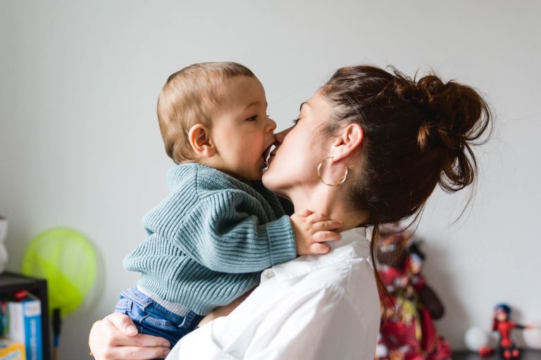 Une femme aux cheveux noirs coiffés en chignon, portant des créoles et une chemise blanche, tient un bébé vêtu d'un pull en tricot vert près de son visage. Le bébé semble toucher le nez ou la bouche de la femme avec sa bouche. Pour capturer ce moment de tendresse, l'arrière-plan révèle des jouets et une étagère avec des livres, parfaits pour leur séance photo de famille Bordelaise.